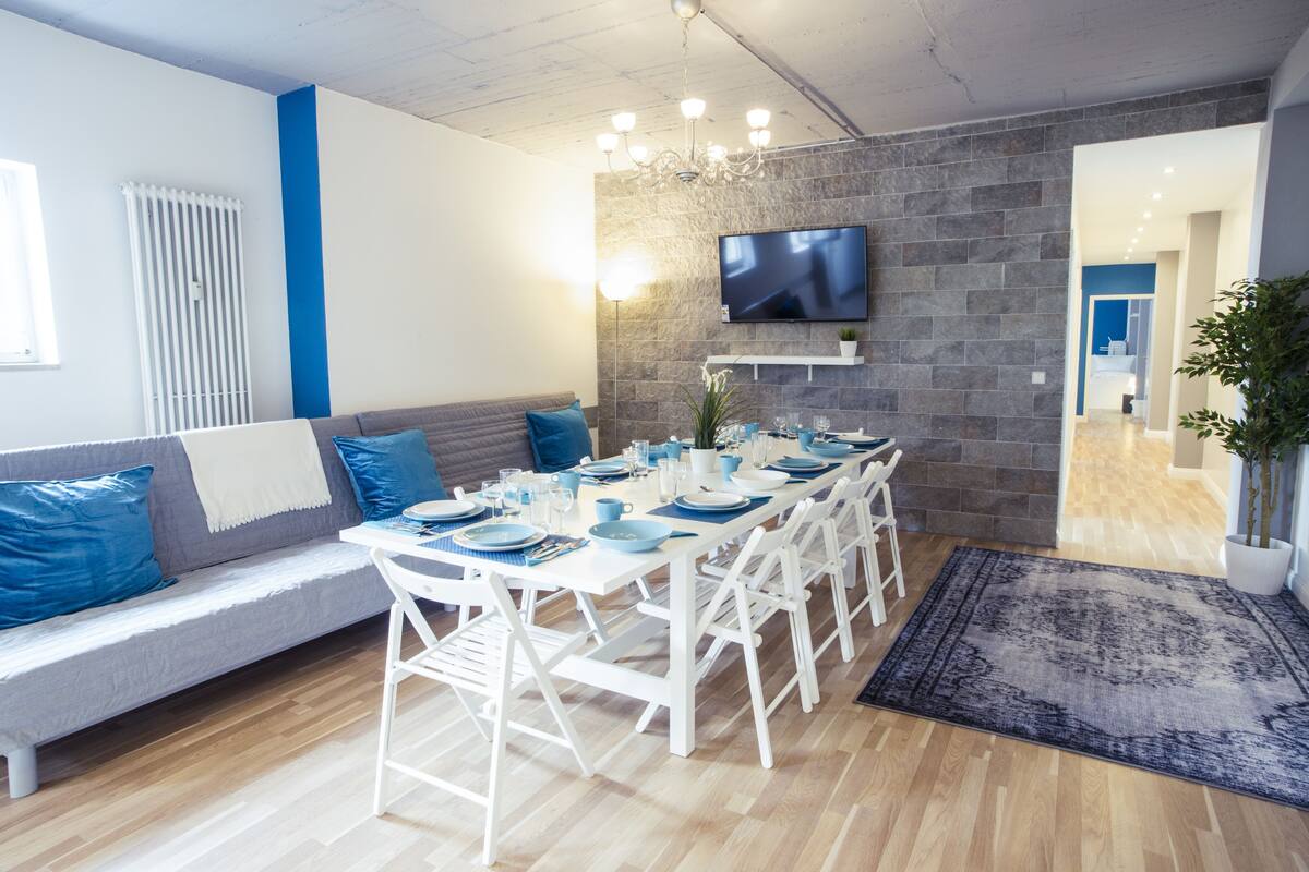 Un espace de salle à manger moderne avec une longue table dressée pour six personnes, des chaises blanches, un canapé gris avec des coussins bleus, un téléviseur fixé au mur et un mur en pierre.