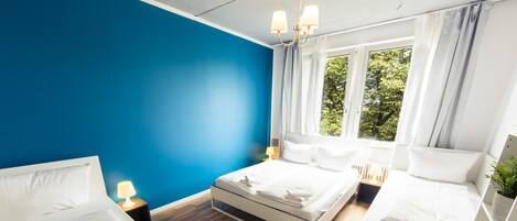 A modern hotel room with two beds, a blue accent wall, and large windows with curtains.