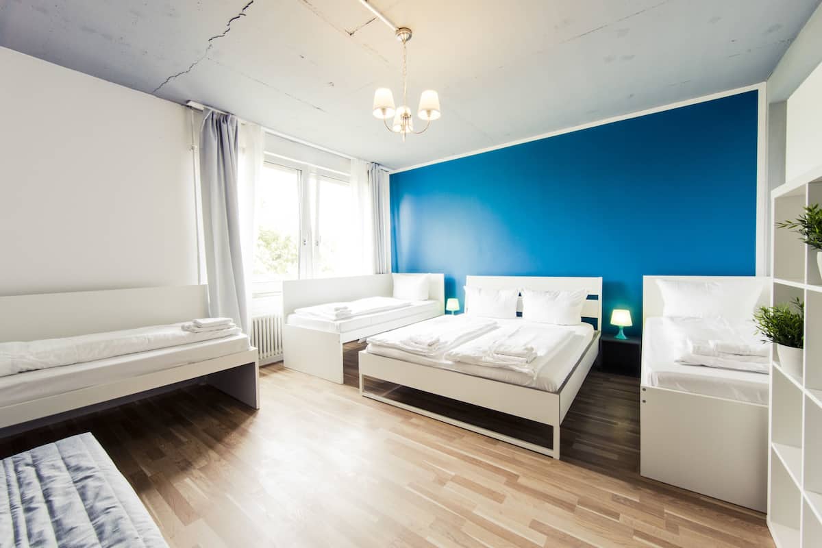 Une chambre d’hôtel moderne avec un plancher en bois, un grand lit, un plus petit lit, un banc, une table de chevet avec une lampe et une plante.
