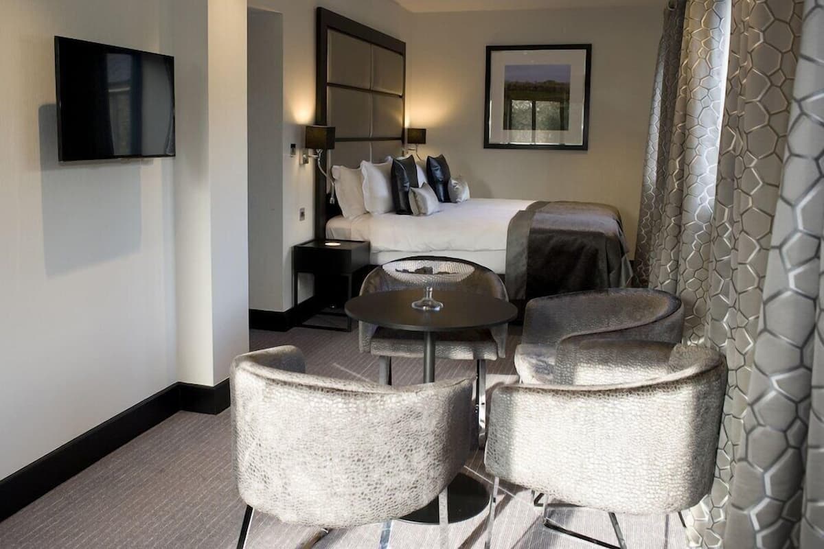 A hotel room with a bed, a TV, a round table, and two armchairs.