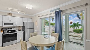 A kitchen with white cabinets, a stainless steel refrigerator, and a microwave. There is a dining table with chairs and a view of the beach through the glass door.