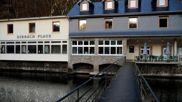 A riverside building with a stone bridge, named DIRBACH PLAGE, featuring multiple windows and a covered outdoor seating area. - Hotel Dirbach Plage (Esch-sur-Sure)