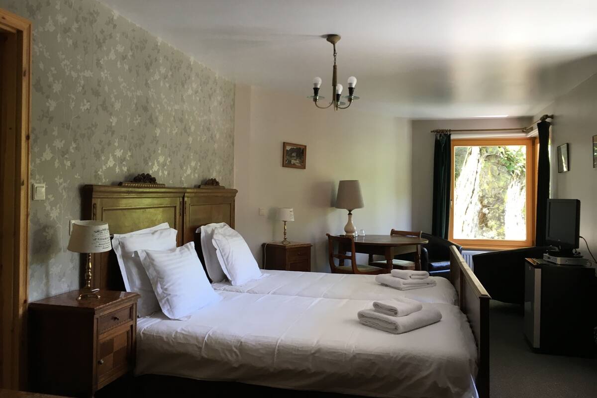 A hotel room with a bed, bedside tables, a desk, a chair, a television, and a window with a view of trees.