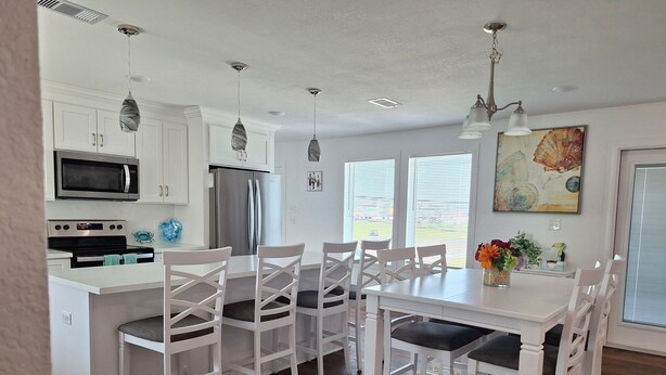 A modern kitchen with white cabinetry, stainless steel appliances, and a dining area with white chairs and a table. - Kids Cottage Amazing views from all directions (Jamaica Beach)