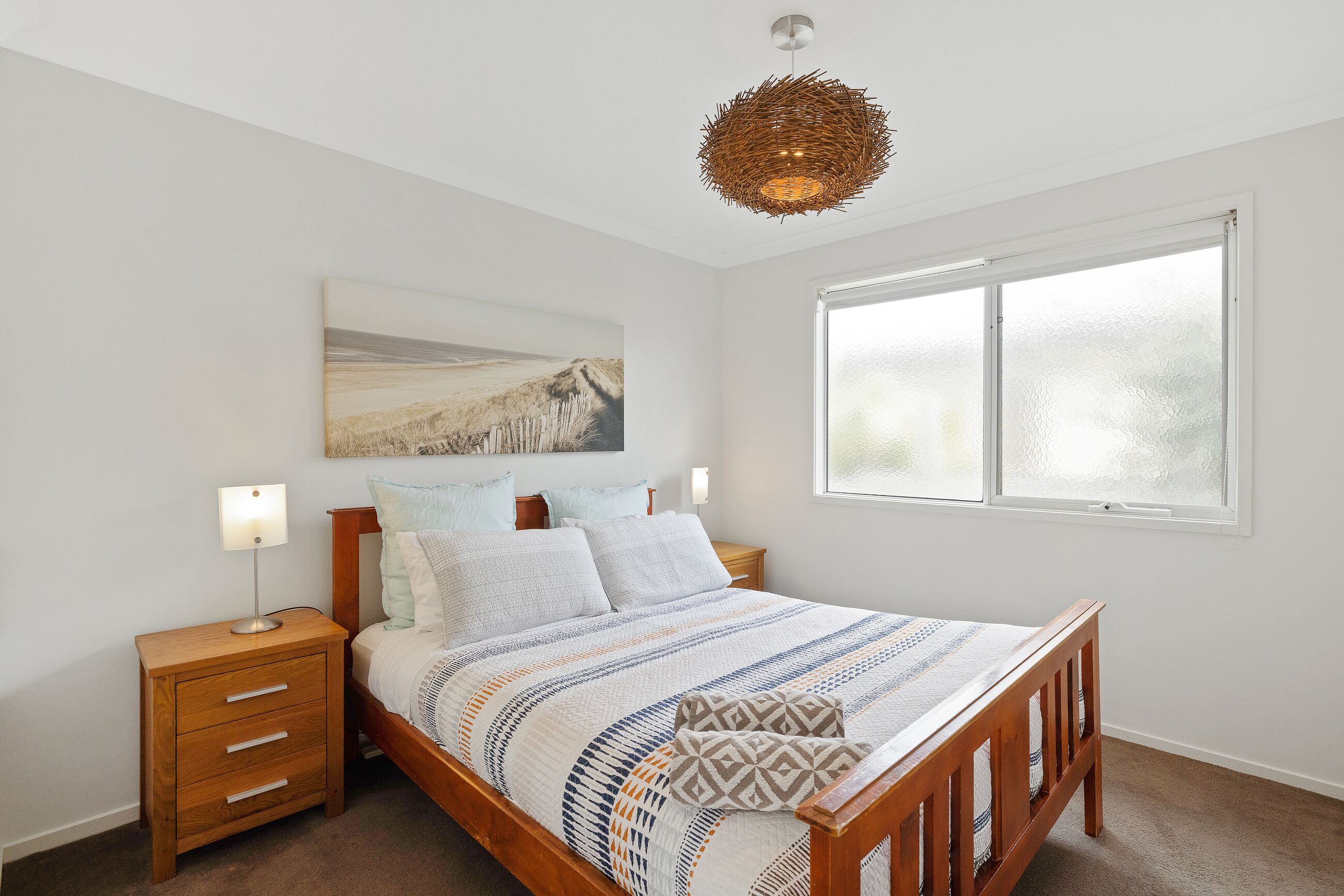 Une chambre avec un lit en bois, une table de chevet avec des tiroirs, une lampe fixée au mur, une grande fenêtre et un luminaire suspendu.