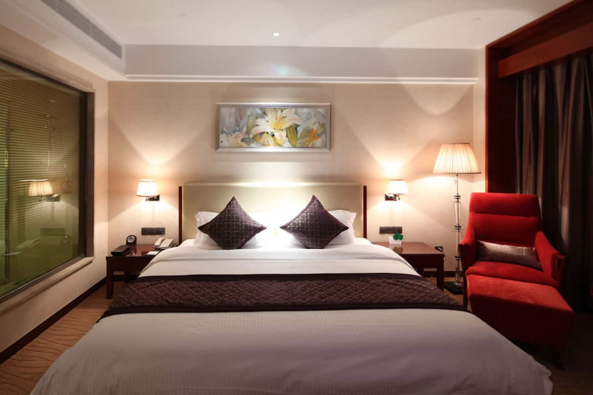 Een hotelkamer met een groot bed, twee bedlampen, een schilderij aan de muur, een rode fauteuil en een raam met zonwering.