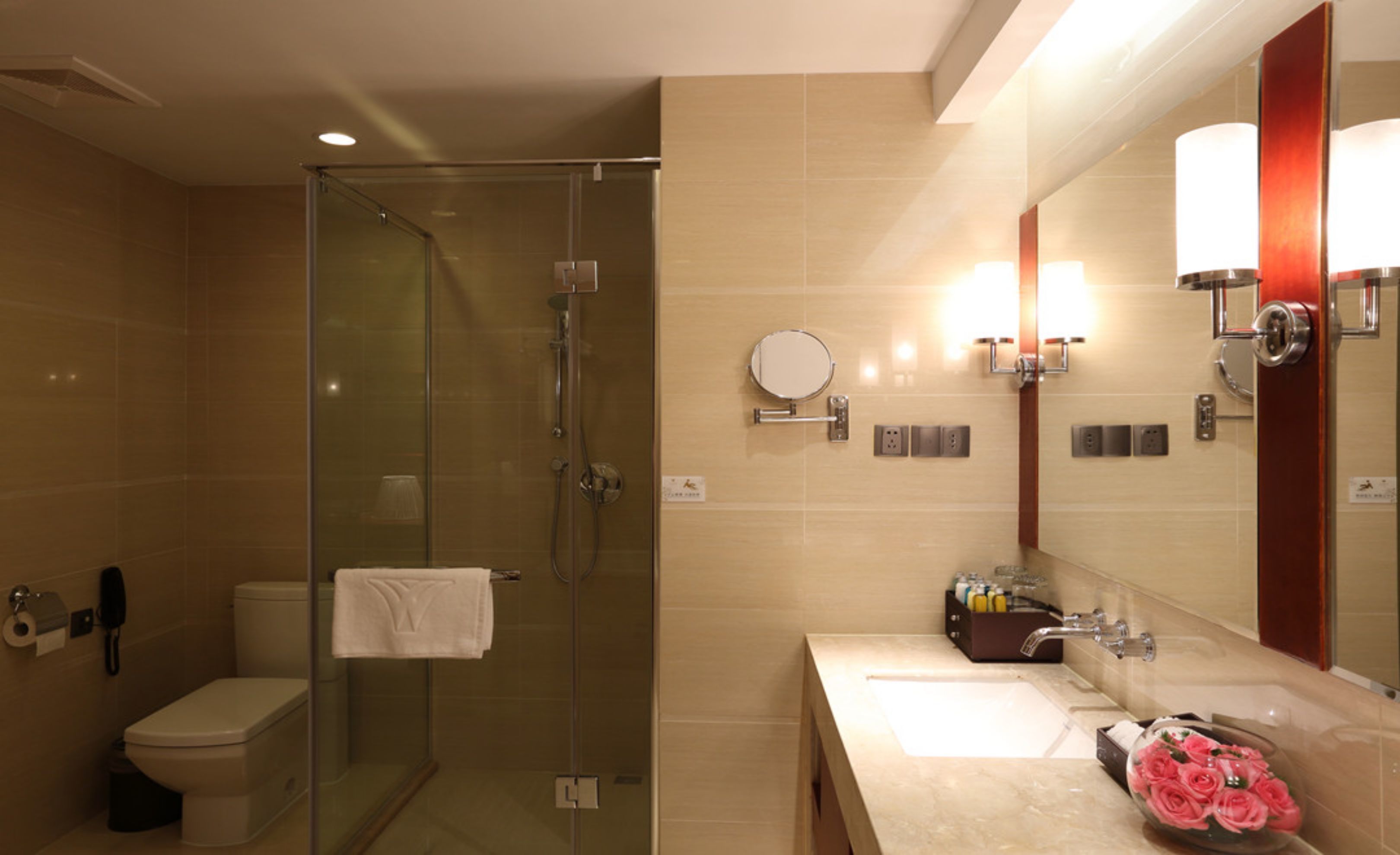 A modern bathroom with a glass-enclosed shower, a toilet, a sink with a mirror, and a vase of pink flowers.