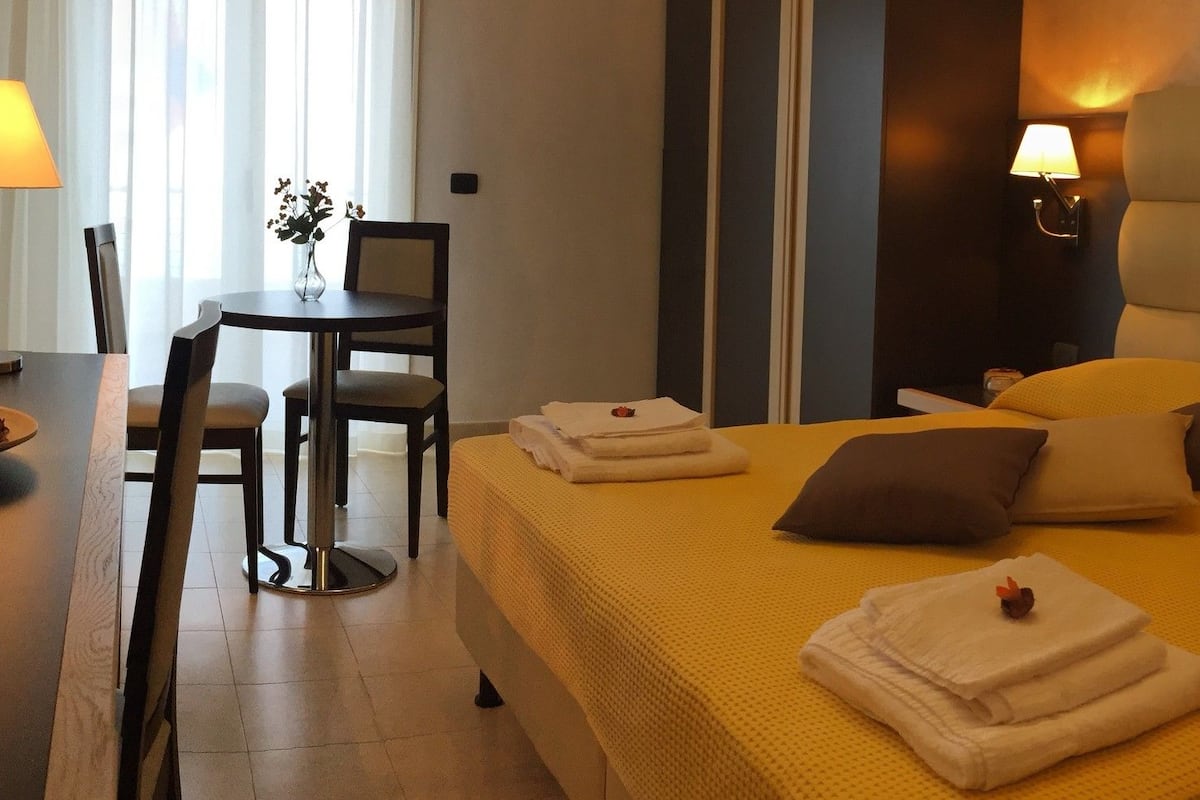 Camera d'albergo con un letto, comodini, una sedia e un tavolino con un vaso e una ciotola.