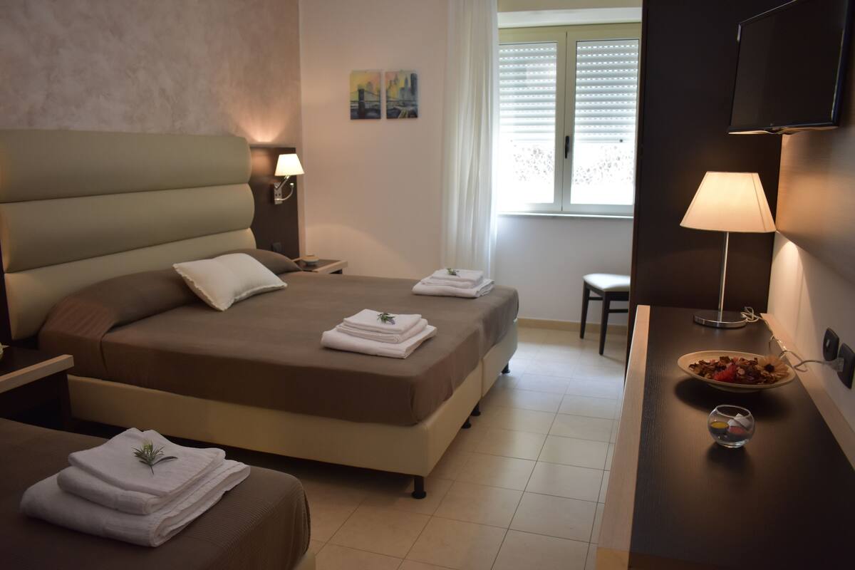 Camera d'albergo con un letto grande, un comodino, una scrivania con una lampada, una sedia e una finestra con persiane.