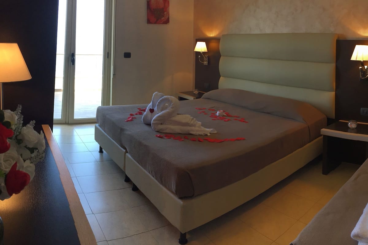 Una camera d'albergo con un letto grande, un letto più piccolo, un comodino, una lampada e un vaso di fiori.