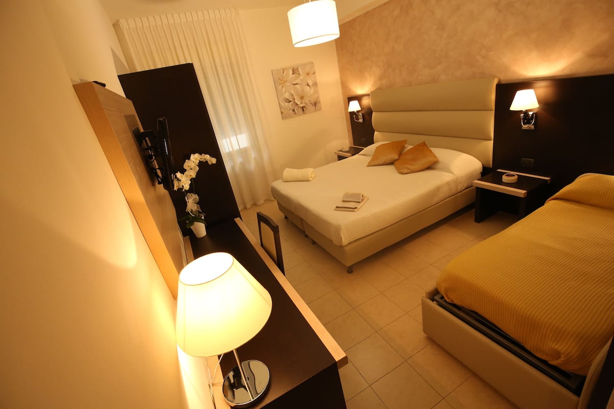 Una camera d'albergo con un letto, comodini, una scrivania, una sedia, una lampada, un portafoto e un vaso con fiori.