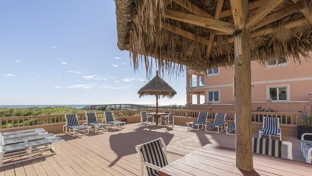 A wooden deck with striped lounge chairs and a thatched umbrella, overlooking a beach and a pink building. - Suntide II 301 - Oceanfront Condo, BBQ's, Sand Volleyball, Sun Deck, Beachfront Pool & Spa (South Padre Island)