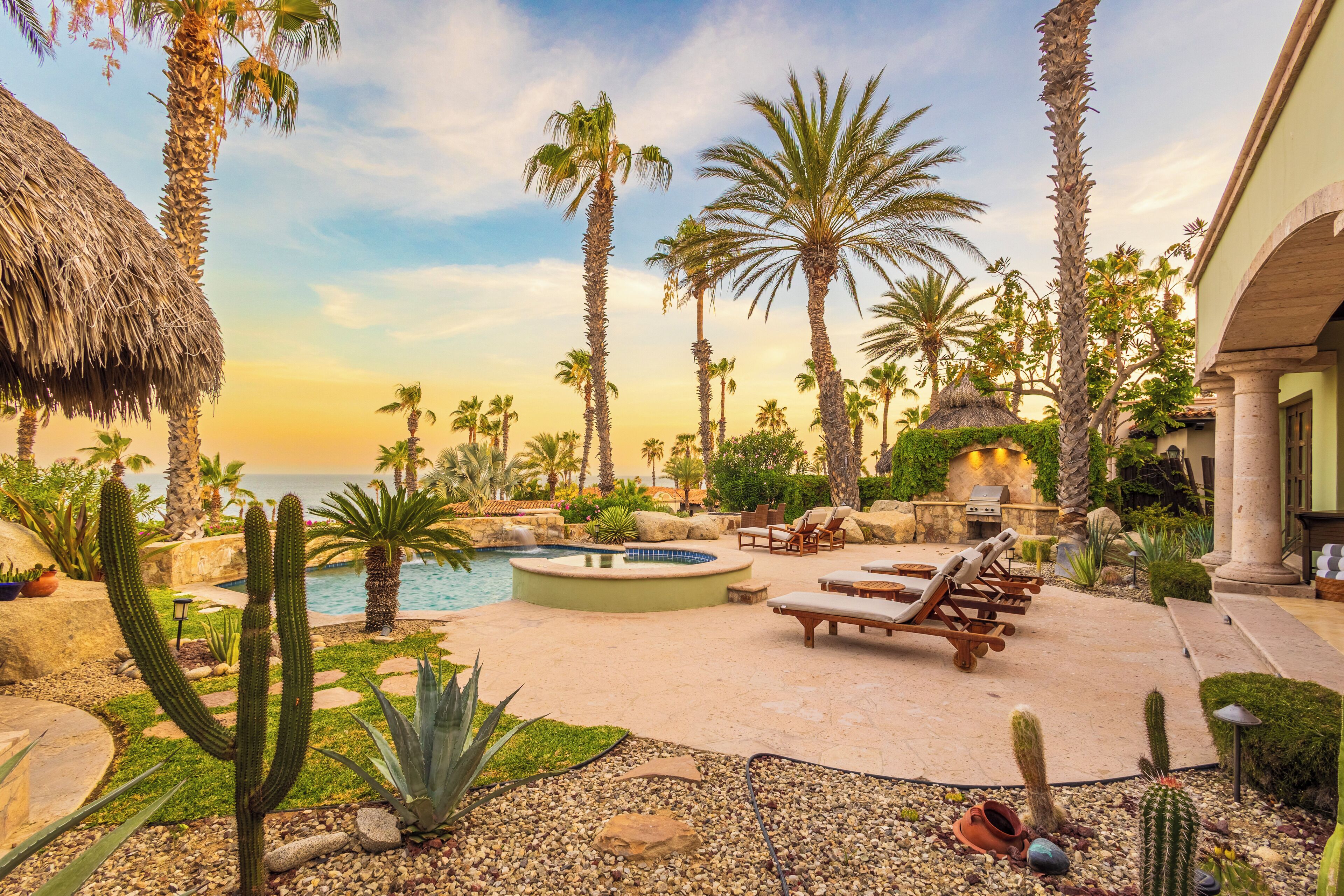 A pool area with lounge chairs, palm trees, and a thatched roof structure.