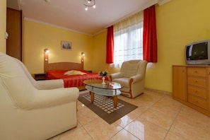 A hotel room with a bed, a sofa, a glass coffee table, a TV, and a wooden cabinet. - Aqua Blue Hotel (Hajduszoboszlo)