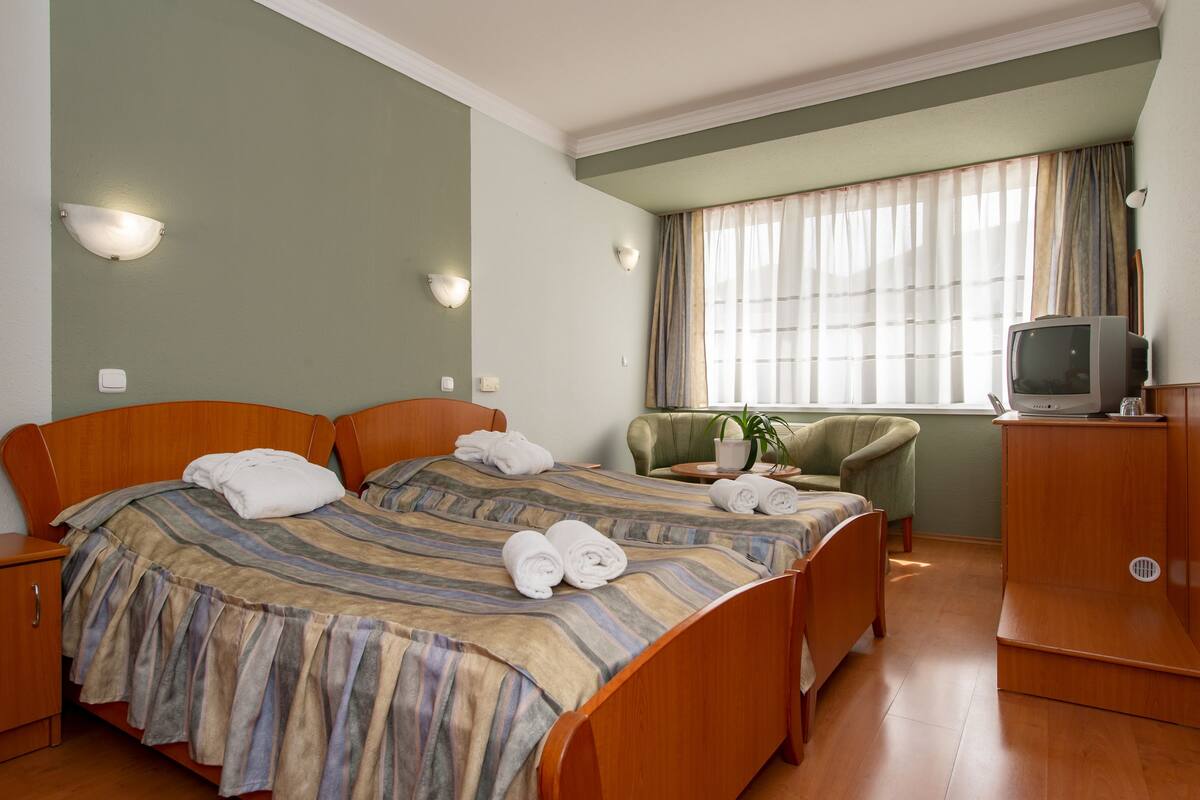Ein Hotelzimmer mit zwei Betten, einem Fernseher, einem Schreibtisch und einem Fenster mit Vorhängen.