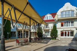 A hotel courtyard with outdoor seating, a covered area, and a building with a red roof and white facade. - Aqua Blue Hotel (Hajduszoboszlo)