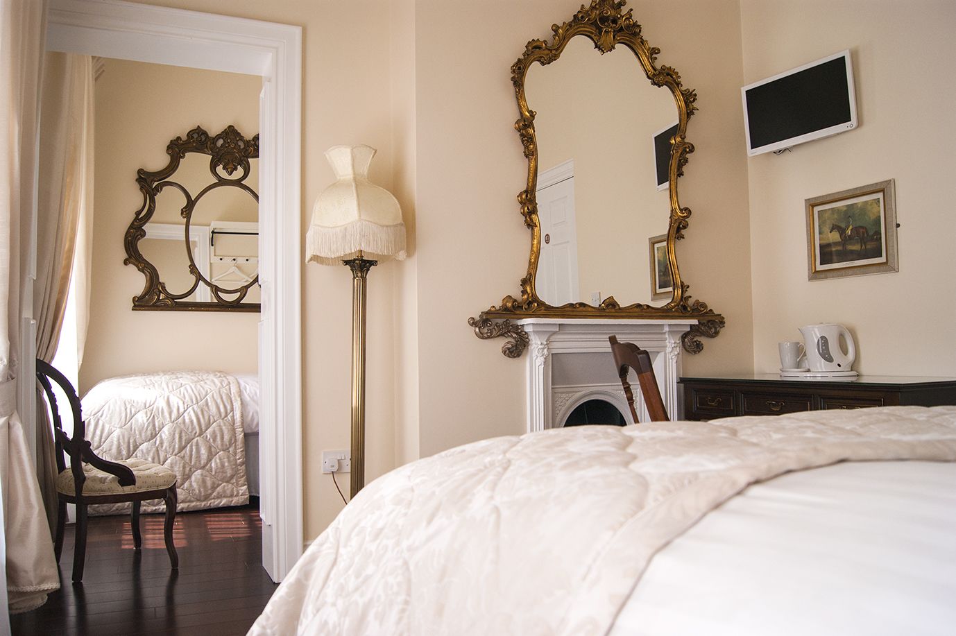 A hotel room with a large bed, a sitting area with a chair, a fireplace, and two ornate mirrors.