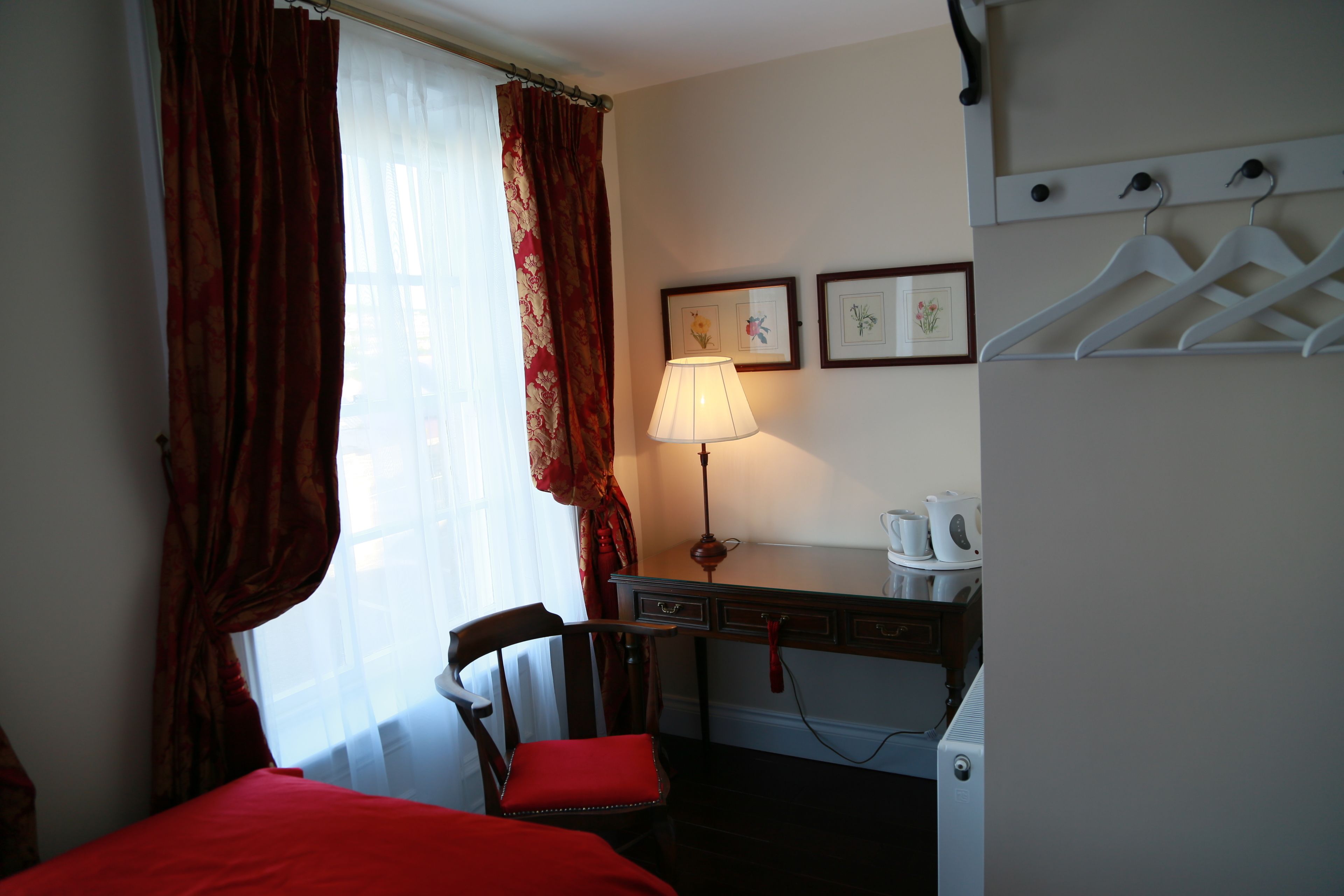 Habitación de hotel con cama, una silla, un escritorio con lámpara, radiador y perchero con ganchos.