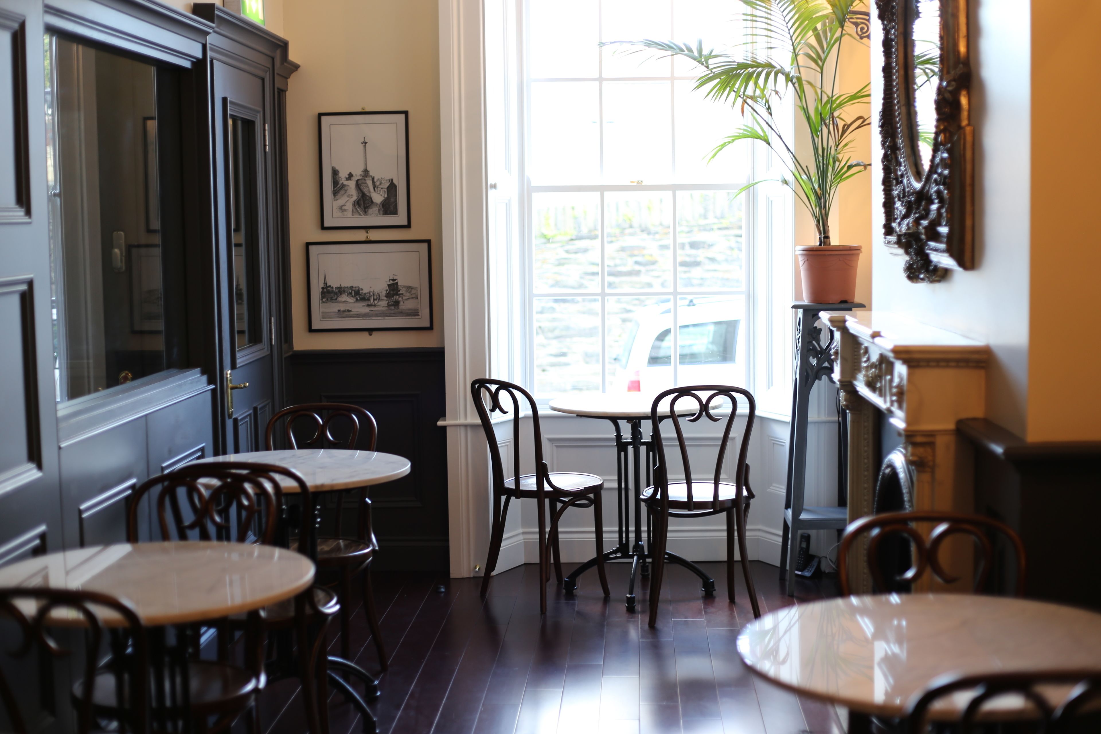 A dining area with wooden tables and chairs, a large window with a view of the street, and framed pictures on the wall.