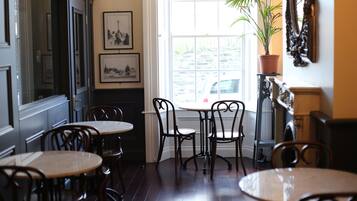 A dining area with wooden tables and chairs, a large window with a view of the street, and framed pictures on the wall.