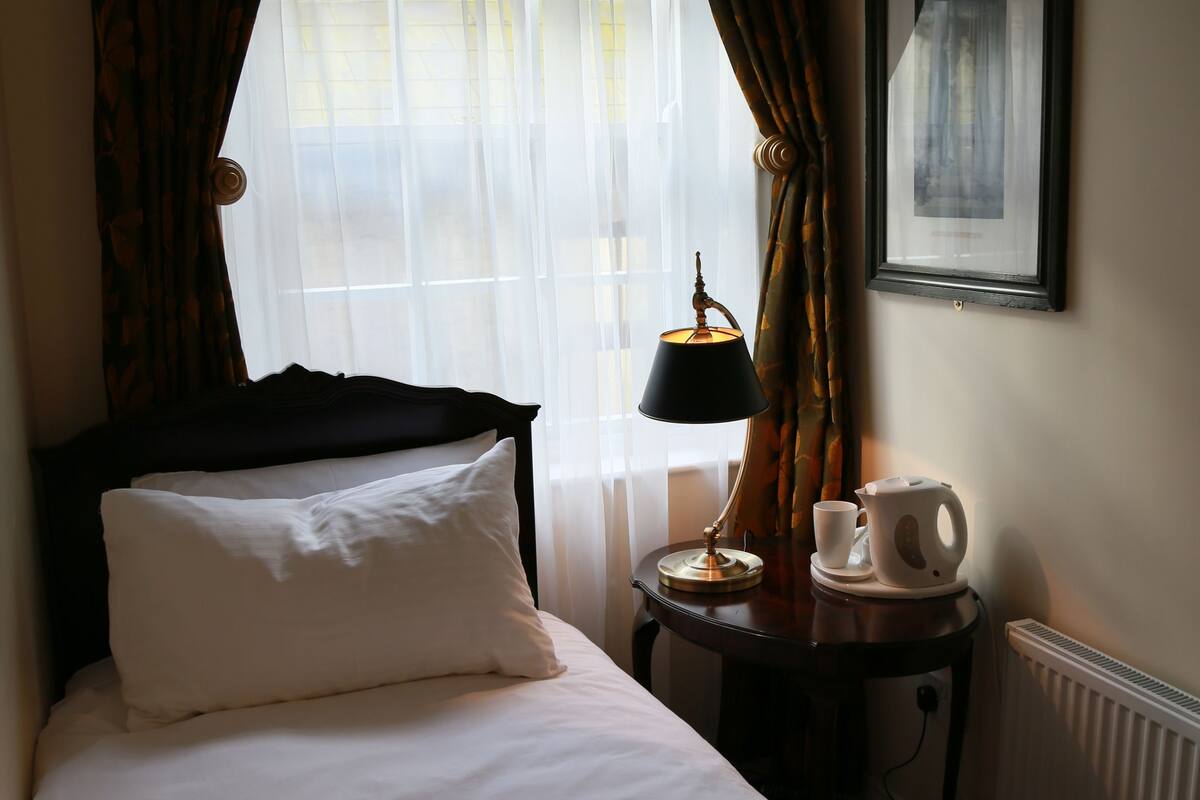 Ein Hotelzimmer mit Bett, Nachttisch mit Lampe, gerahmtes Bild an der Wand und Fenster mit Vorhängen.