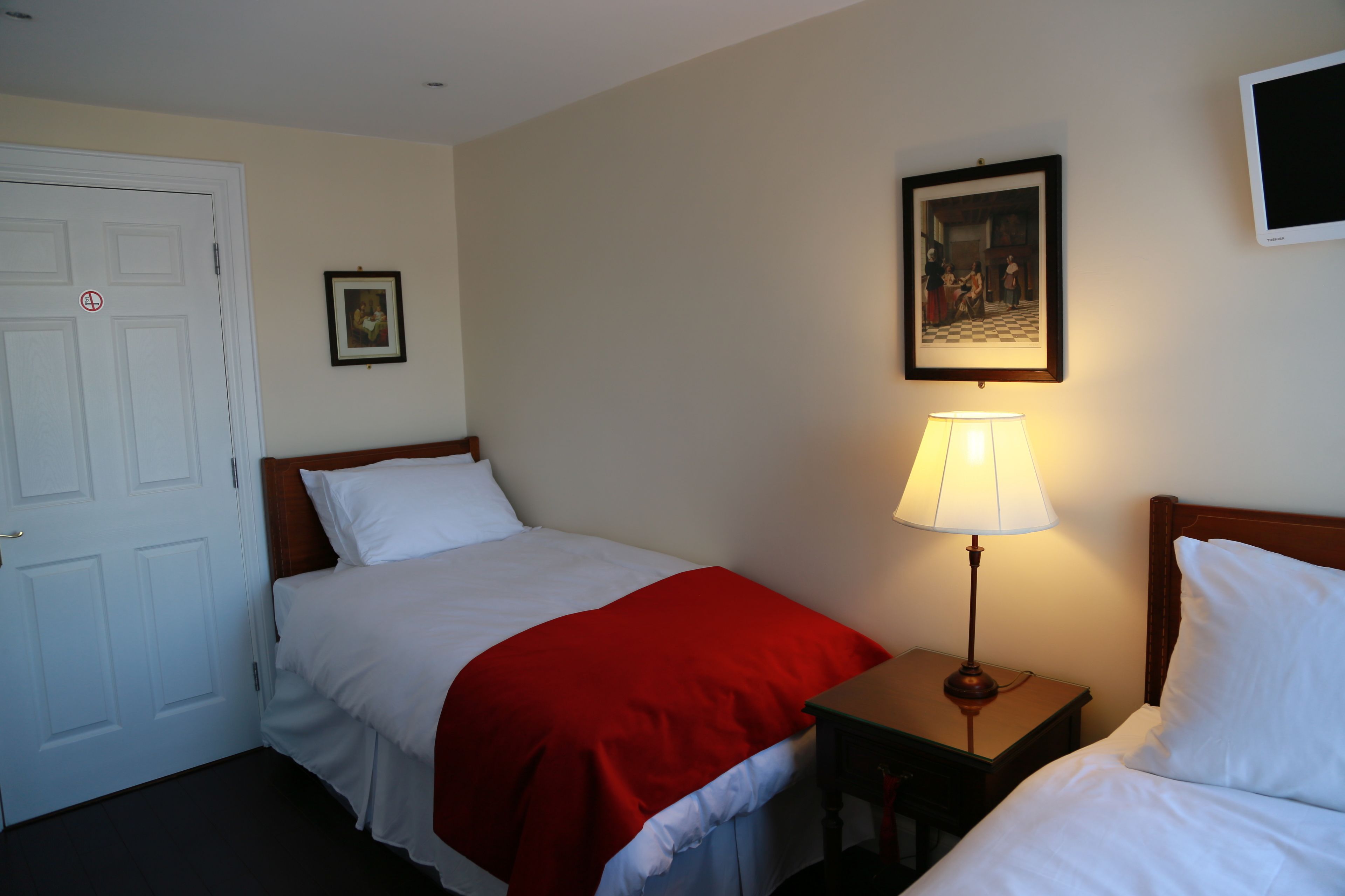 A hotel room with two single beds, a nightstand with a lamp, a door with a no entry sign, and a framed picture on the wall.