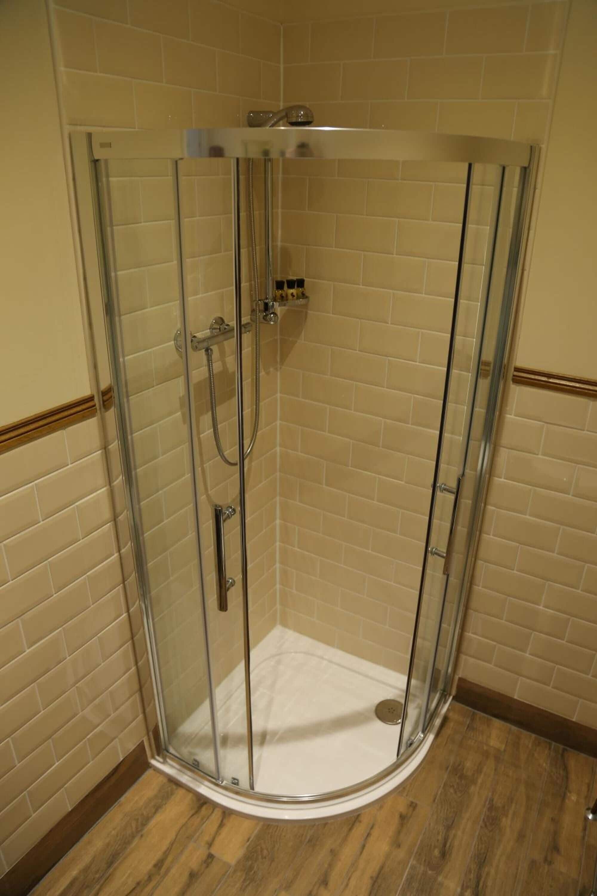 A corner shower with a glass door, beige tiled walls, and wooden flooring.