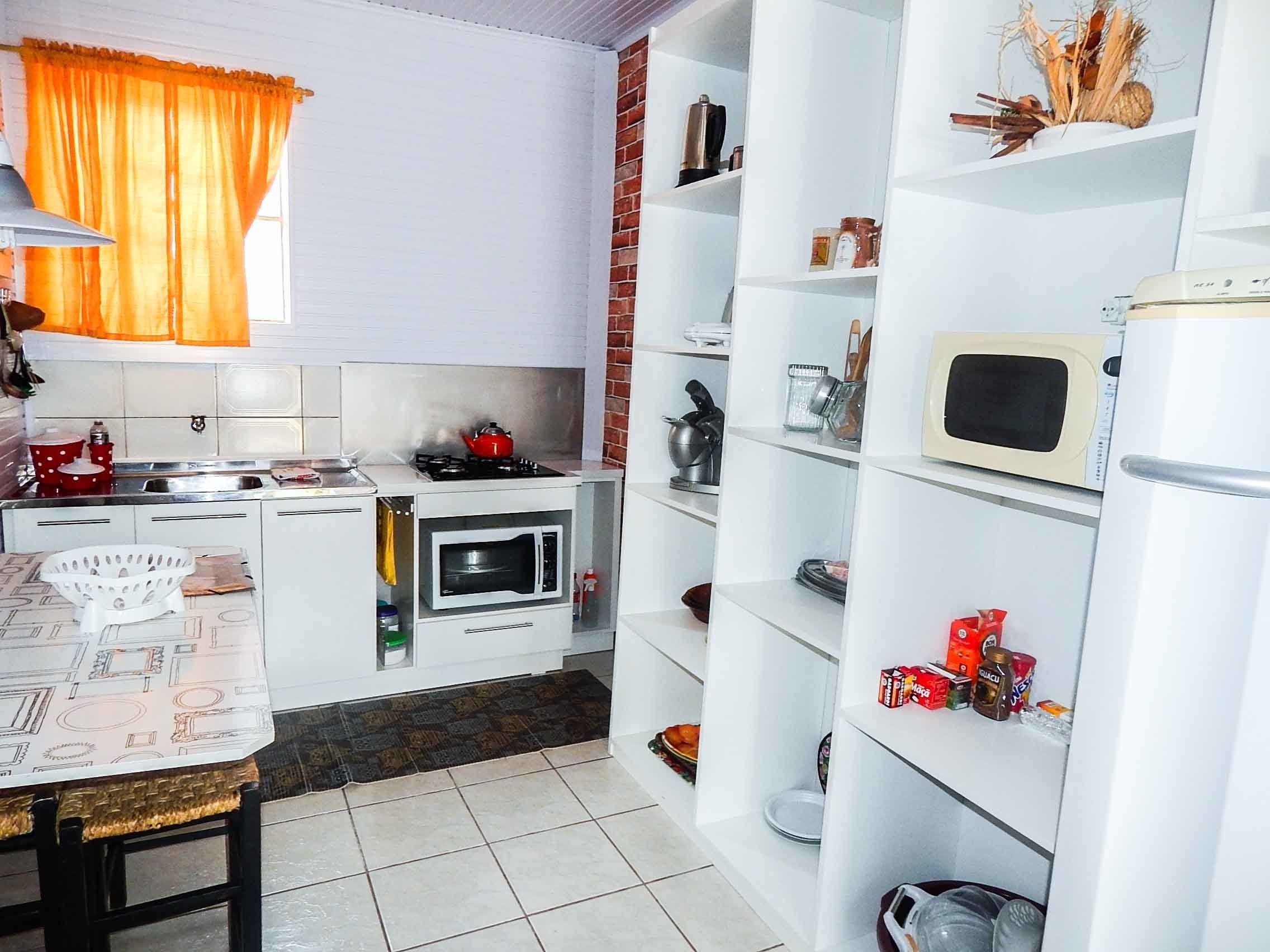 A compact kitchen with a gas stove, microwave, and a small oven. There is a dining table with a basket and a chair. The kitchen has white cabinets and a brick wall.