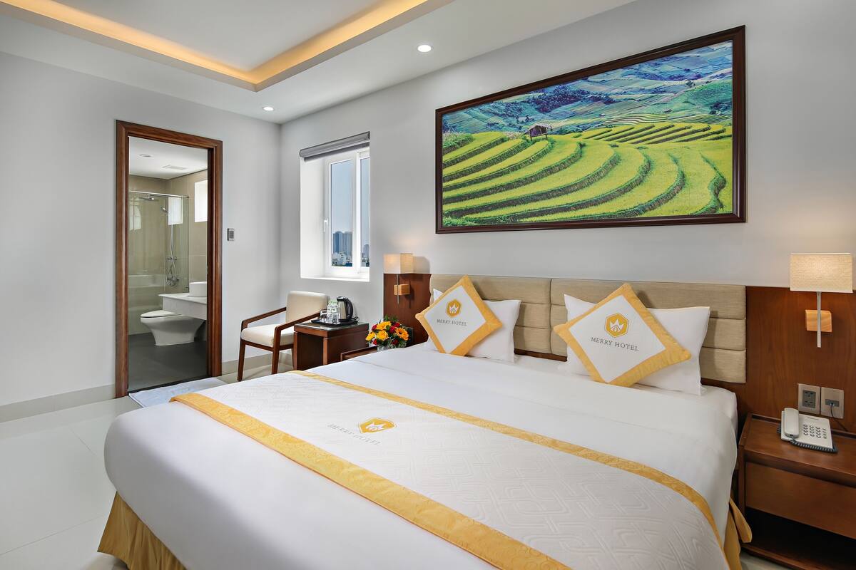 A hotel room with a large bed, a painting of a landscape, a desk with a lamp, and a bathroom visible through an open door.