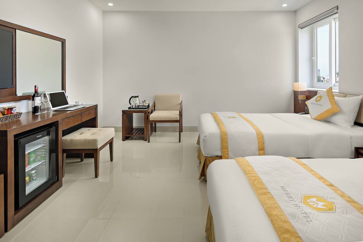 A hotel room with a minibar, a desk with a laptop, a chair, two beds with matching headboards, and a large window.