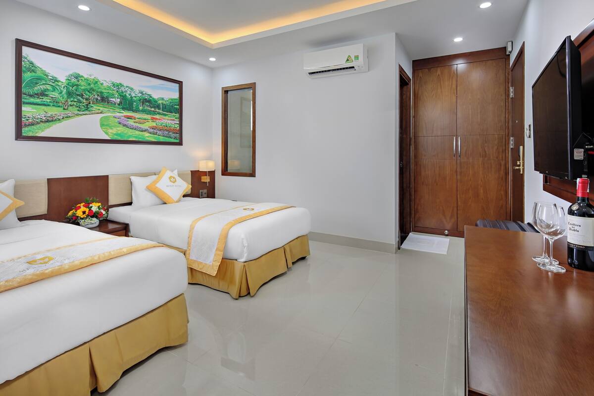 A hotel room with two beds, a large painting of a landscape, an air conditioning unit, a wardrobe, a television, and a table with a bottle and glass.