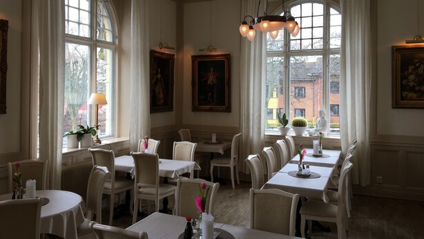 A dining area with multiple tables and chairs, large windows with curtains, and paintings on the walls.