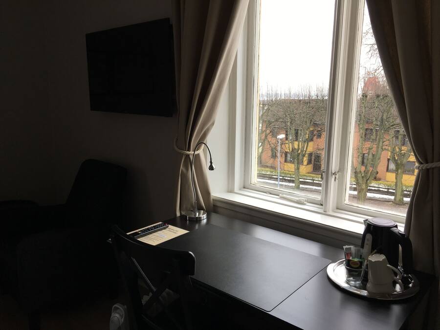 Habitación de hotel con escritorio, una silla, ventana con cortinas y vistas a edificios. Hay un hervidor sobre la mesita.