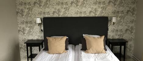 Una cama doble con cabecera oscura, ropa de cama blanca y dos almohadas color beige. Hay dos mesitas de noche con lámparas a cada lado. La pared tiene un papel tapiz con motivos de paisaje natural.