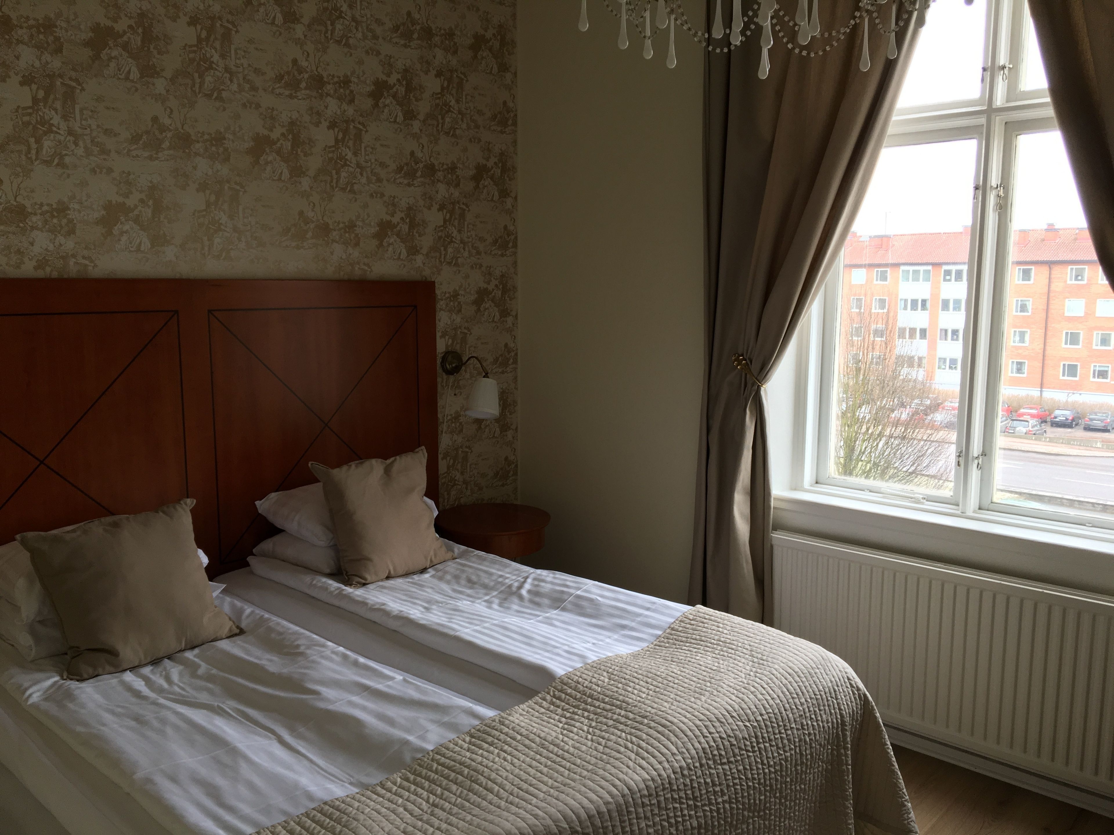 Un dormitorio con cabecera de madera, cama con ropa blanca, colcha color beige y vistas a un edificio a través de una ventana con cortinas.