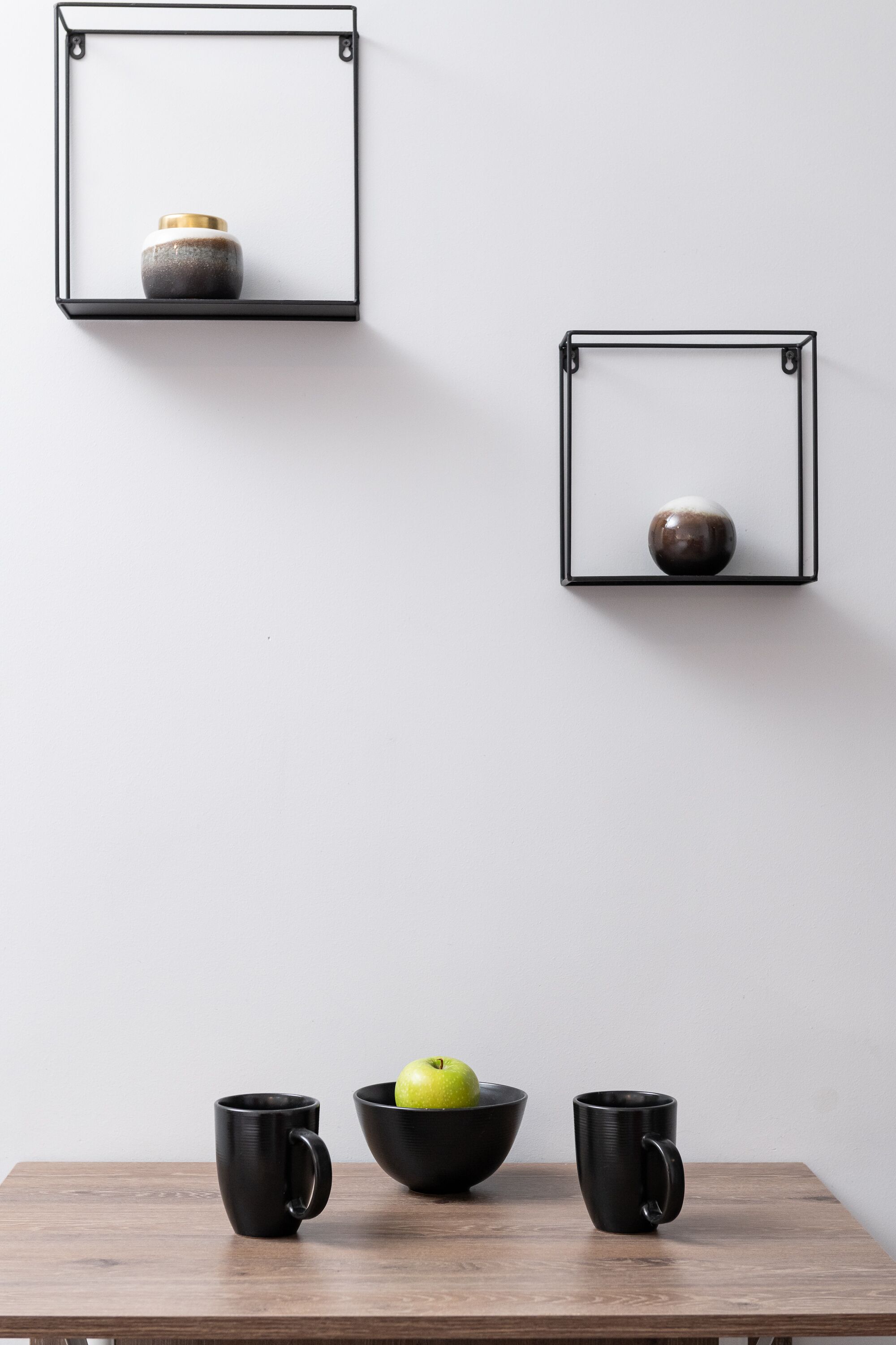 A minimalist room with a wooden table, two black mugs, a bowl with an apple, and two wall-mounted shelves with decorative items.