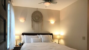 Un lit bien fait, avec du linge de lit blanc, un attrape-rêves accroché au mur, deux lampes de chevet et un ventilateur de plafond.