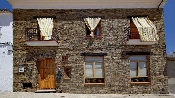 A stone building with a wooden door, windows with shutters, and a balcony with striped curtains. - Casa Rural La Breña (Talavan)
