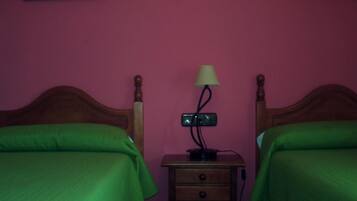Een kamer met twee houten bedden, elk met een groene plaid, een nachtkastje met een lamp, en een wandcontactdoos tussen de bedden.