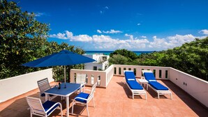 Eine Terrasse mit Tisch und Stühlen, einem blauen Sonnenschirm und Liegestühlen mit blauen Kissen.