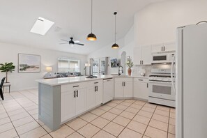 Een moderne keuken met witte kasten, een centraal eiland, een plafondventilator, hanglampen, een magnetron, een oven en een koelkast.
