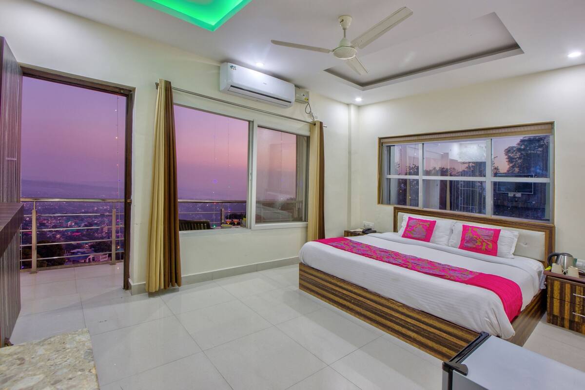 A modern hotel room with a large bed, a balcony with a city view, and an air conditioning unit on the wall.
