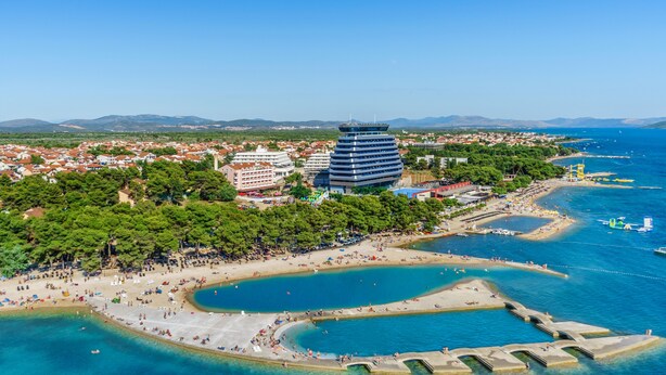 Vista aèria d'una ciutat costanera amb un edifici modern destacat, una platja i aigües blaves cristal·lines.