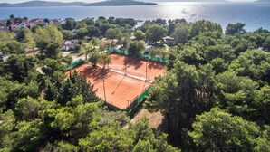 Vista aèria d'una zona costera amb una pista de tennis envoltada per arbres densos i edificis residencials.