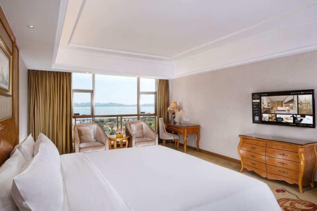 A hotel room with a large bed, a desk, a chair, a TV, and a view of the sea.