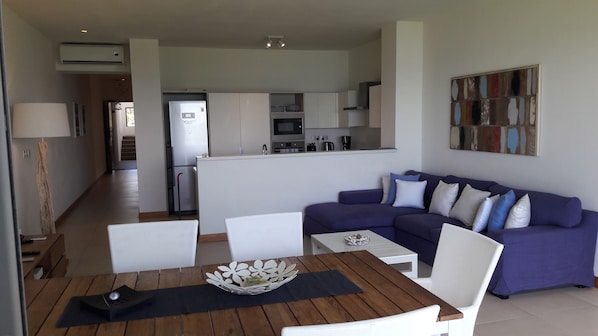 Una sala de estar moderna con un sofá morado, sillones blancos y una mesa de centro de madera. Se ve una cocina al fondo, con armarios blancos y un microondas.