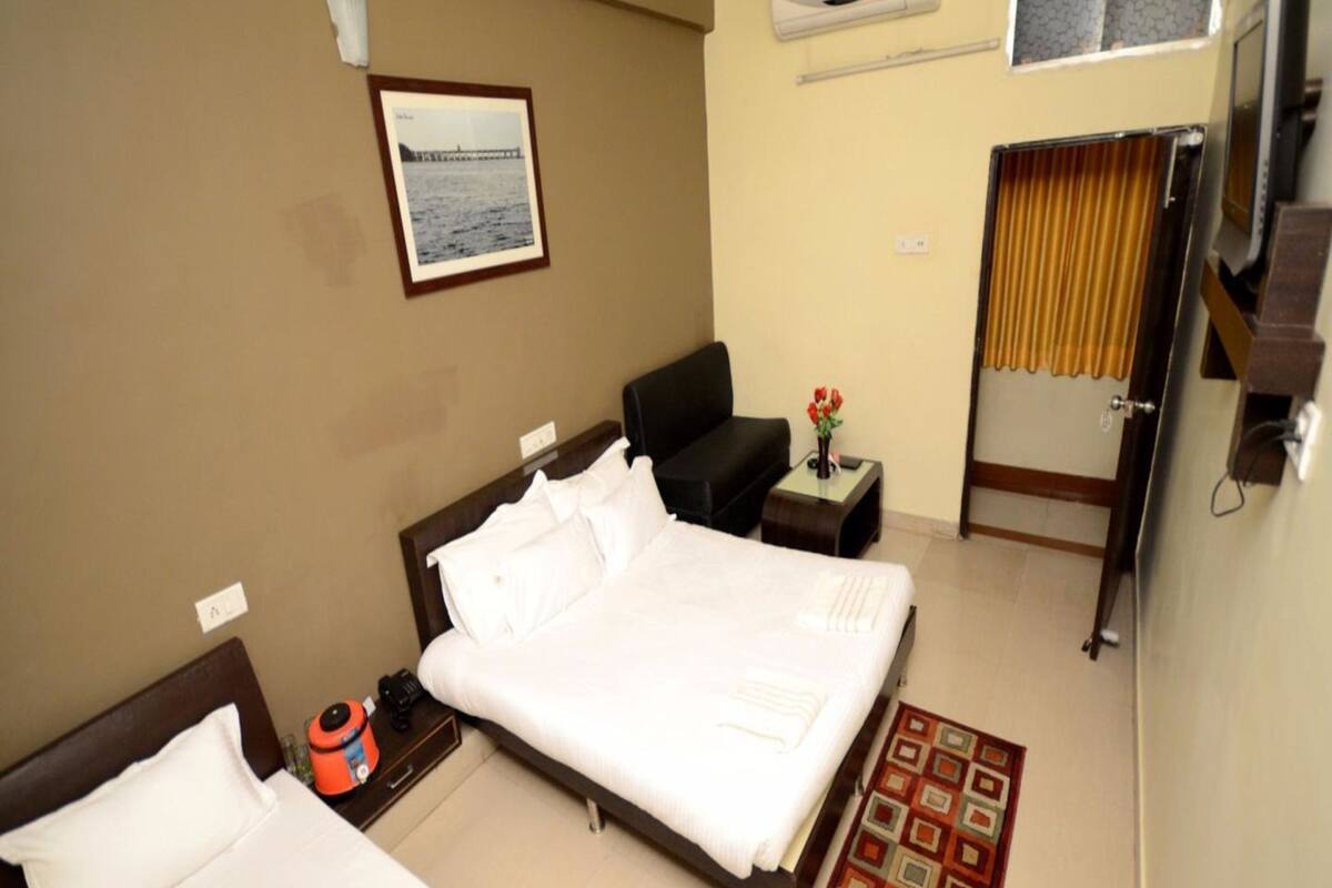 A hotel room with a large bed, a smaller bed, a desk with a chair, an air conditioner, a framed picture on the wall, and a window with curtains.