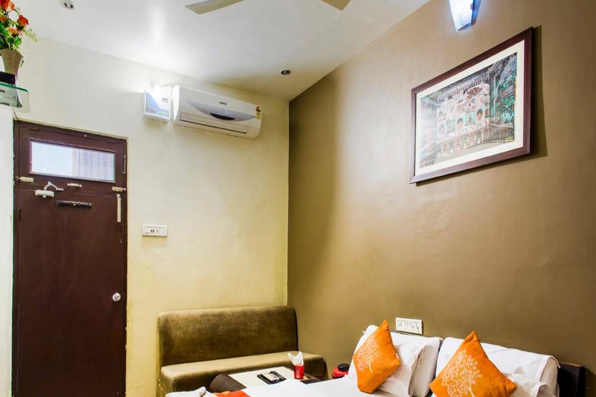 A hotel room with a bed, a sofa, an air conditioning unit, a framed picture on the wall, and a door.