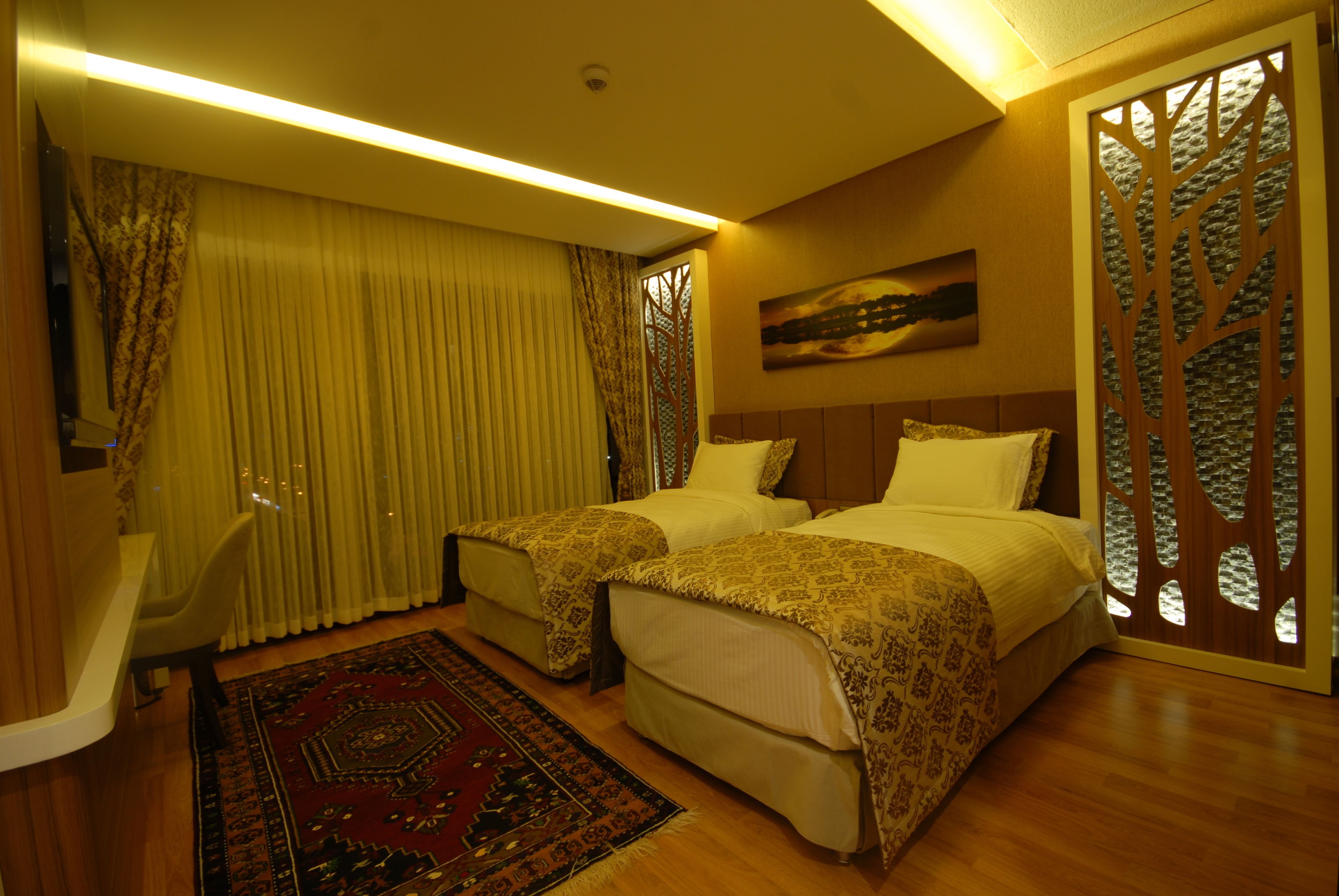 Habitación de hotel con dos camas, un escritorio, una silla, un ventanal con cortinas, un panel decorativo en la pared y una alfombra.