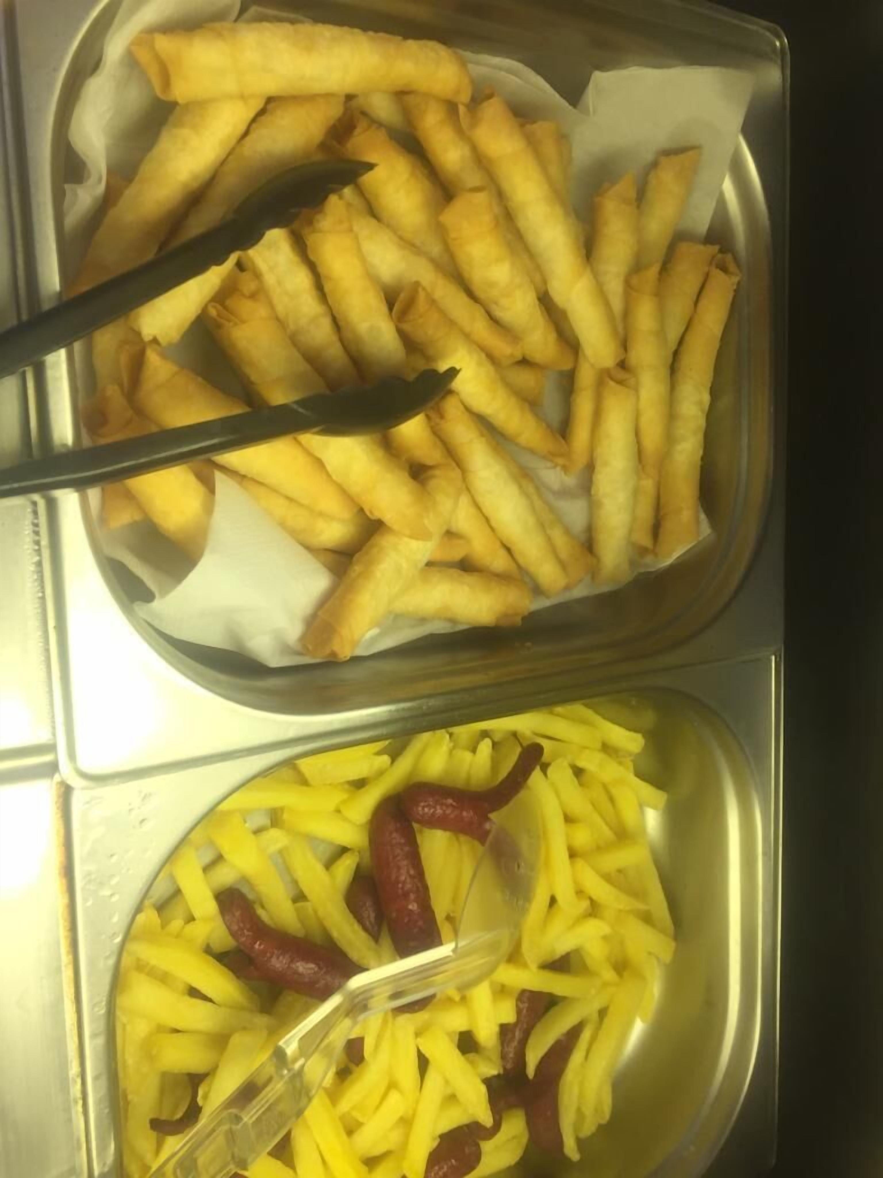 Uma bandeja com rolinhos primavera dourados e caseiros e um recipiente com batatas fritas, acompanhado de um garfo de plástico.